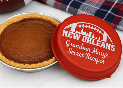 &quot;Personalized red pie tin lid engraved with a football skyline design and the text &#39;New Orleans – Grandma Mary’s Secret Recipes.&#39; The lid rests beside a freshly baked pie on a marble countertop, with a plaid towel and football in the background. Text at the bottom reads &#39;Baked with love.&#39;&quot;