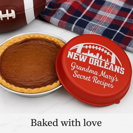 "Personalized red pie tin lid engraved with a football skyline design and the text 'New Orleans – Grandma Mary’s Secret Recipes.' The lid rests beside a freshly baked pie on a marble countertop, with a plaid towel and football in the background. Text at the bottom reads 'Baked with love.'"
