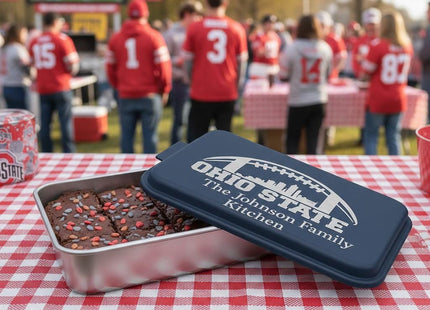 &quot;Personalized navy blue aluminum cake pan engraved with &#39;Ohio State – The Johnson Family Kitchen&#39; sitting on a red and white checkered table at an outdoor football tailgate. The pan contains brownies topped with red and white sprinkles. In the background, fans wearing red jerseys watch a football game on a large screen under sunny skies.&quot;