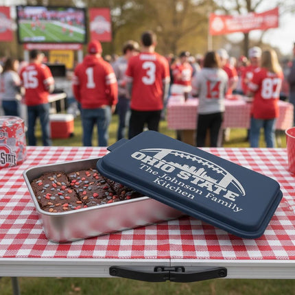 "Personalized navy blue aluminum cake pan engraved with 'Ohio State – The Johnson Family Kitchen' sitting on a red and white checkered table at an outdoor football tailgate. The pan contains brownies topped with red and white sprinkles. In the background, fans wearing red jerseys watch a football game on a large screen under sunny skies."