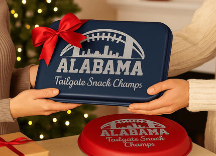 &quot;Two people exchange a personalized Alabama football-themed bakeware gift set in a warm holiday setting. One holds a navy blue aluminum cake pan with a red bow, engraved with &#39;Alabama Tailgate Snack Champs,&#39; while a red pie tin with the same engraving rests on the table beside a wrapped present. A Christmas tree and twinkling lights create a festive background.&quot;