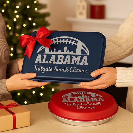 "Two people exchange a personalized Alabama football-themed bakeware gift set in a warm holiday setting. One holds a navy blue aluminum cake pan with a red bow, engraved with 'Alabama Tailgate Snack Champs,' while a red pie tin with the same engraving rests on the table beside a wrapped present. A Christmas tree and twinkling lights create a festive background."