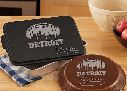 &quot;Personalized Detroit football-themed bakeware set displayed on a wooden kitchen counter. The set includes a black aluminum cake pan and a brown pie tin, both laser engraved with a football design, the Detroit skyline, and the name &#39;Peterson&#39; with &#39;Darin & Tony&#39; below. A blue and white checkered towel, mixing bowls, and apples complete the cozy kitchen scene.&quot;