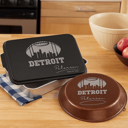 "Personalized Detroit football-themed bakeware set displayed on a wooden kitchen counter. The set includes a black aluminum cake pan and a brown pie tin, both laser engraved with a football design, the Detroit skyline, and the name 'Peterson' with 'Darin & Tony' below. A blue and white checkered towel, mixing bowls, and apples complete the cozy kitchen scene."