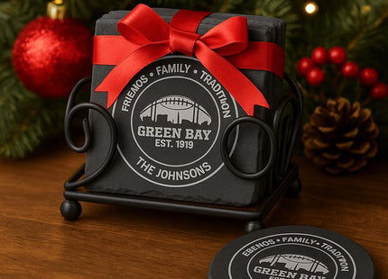 &quot;Personalized Green Bay slate coasters engraved with &#39;Friends • Family • Tradition,&#39; &#39;Green Bay Est. 1919,&#39; and &#39;The Johnsons,&#39; displayed in a black metal holder tied with a red satin bow. Set on a wooden table with Christmas decorations including a pine tree, red ornaments, and pinecones in the background.&quot;