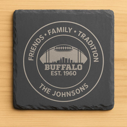 "Square black slate coaster engraved with a Buffalo football design featuring the text 'Friends • Family • Tradition,' 'Buffalo Est. 1960,' and 'The Johnsons.' The engraving shows a football silhouette with a city skyline inside, displayed on a light wood background."