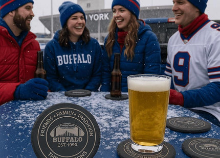 &quot;Four friends dressed in blue and red winter gear enjoying a snowy tailgate party outside a stadium. They sit around a blue table with beer bottles and a pint of beer on personalized Buffalo football coasters engraved with &#39;Friends • Family • Tradition,&#39; &#39;Buffalo Est. 1990,&#39; and &#39;The Johnsons.&#39; Snow lightly covers the table and coasters, creating a festive game-day atmosphere.&quot;