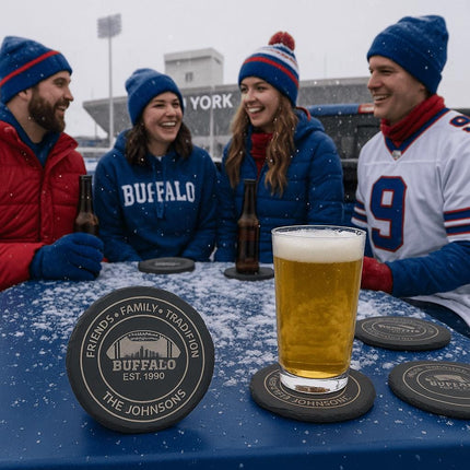 "Four friends dressed in blue and red winter gear enjoying a snowy tailgate party outside a stadium. They sit around a blue table with beer bottles and a pint of beer on personalized Buffalo football coasters engraved with 'Friends • Family • Tradition,' 'Buffalo Est. 1990,' and 'The Johnsons.' Snow lightly covers the table and coasters, creating a festive game-day atmosphere."