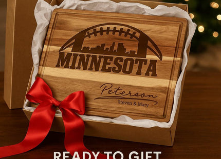&quot;Personalized Minnesota football cutting board made of acacia wood, engraved with skyline and football design, and the name &#39;Peterson – Steven & Mary.&#39; The board is presented in a gift box lined with white tissue paper and tied with a large red satin bow. Text overlay reads &#39;Ready to Gift for Holidays & Housewarmings,&#39; with a softly lit Christmas tree in the background.&quot;