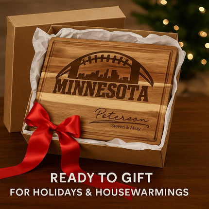 "Personalized Minnesota football cutting board made of acacia wood, engraved with skyline and football design, and the name 'Peterson – Steven & Mary.' The board is presented in a gift box lined with white tissue paper and tied with a large red satin bow. Text overlay reads 'Ready to Gift for Holidays & Housewarmings,' with a softly lit Christmas tree in the background."