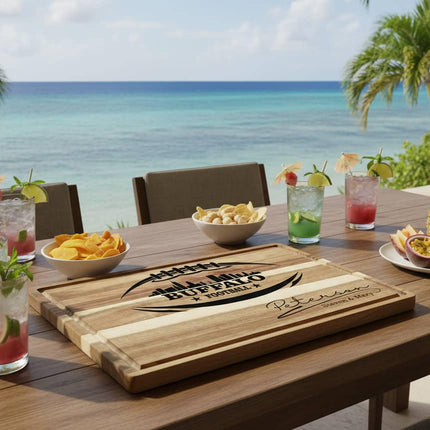 "Personalized Buffalo Football cutting board with engraved football and skyline design and the name 'Peterson – Steven & Mary,' displayed on an outdoor table overlooking a tropical ocean view. The table is set with colorful tropical drinks, bowls of chips and nuts, and a plate of pineapple and passion fruit, creating a relaxed beachside entertaining scene."