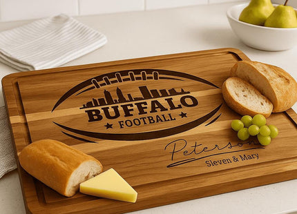 &quot;Personalized Buffalo Football cutting board engraved with football and city skyline design and the name &#39;Peterson – Steven & Mary,&#39; displayed on a kitchen countertop with bread, cheese, and grapes. A white bowl with pears and a folded dish towel are in the background, creating a warm, homey kitchen scene.&quot;