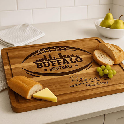 "Personalized Buffalo Football cutting board engraved with football and city skyline design and the name 'Peterson – Steven & Mary,' displayed on a kitchen countertop with bread, cheese, and grapes. A white bowl with pears and a folded dish towel are in the background, creating a warm, homey kitchen scene."