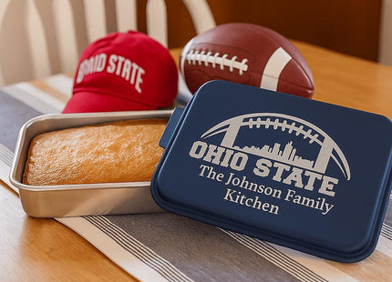 &quot;Personalized navy blue cake pan with the engraving &#39;Ohio State – The Johnson Family Kitchen&#39; featuring a football and city skyline design. The pan sits on a wooden dining table next to a freshly baked cake in the metal base, a red Ohio State cap, and a football, creating a cozy team-themed kitchen scene.&quot;