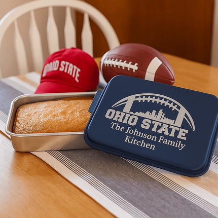"Personalized navy blue cake pan with the engraving 'Ohio State – The Johnson Family Kitchen' featuring a football and city skyline design. The pan sits on a wooden dining table next to a freshly baked cake in the metal base, a red Ohio State cap, and a football, creating a cozy team-themed kitchen scene."