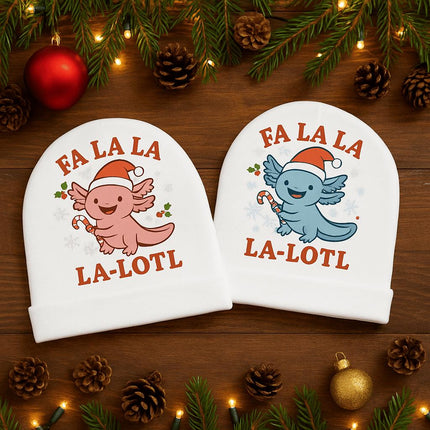 "Two white baby beanies displayed on a wooden surface with holiday decor. One beanie features a pink axolotl wearing a Santa hat and holding a candy cane, with the text 'Fa La La La-Lotl.' The other beanie shows a blue axolotl with the same festive design and text. Pine branches, lights, pinecones, and ornaments surround the beanies in a cozy Christmas setting."