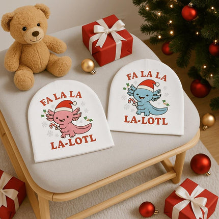 “Two white baby beanies with holiday axolotl designs displayed on a cushioned bench beside a teddy bear and Christmas presents. One beanie features a pink axolotl holding a candy cane with the text ‘FA LA LA LA-LOTL,’ and the other features a blue axolotl with the same design. Christmas ornaments and a decorated tree are visible around the bench.”