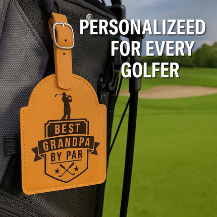 "Tan leather golf bag tag engraved with the phrase 'Best Grandpa by Par' featuring a golfer silhouette and crossed golf clubs, attached to a black and gray golf bag. The bag rests on a lush green golf course with a sand trap in the background. Text at the top reads 'Personalized for Every Golfer.'"