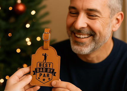 &quot;Smiling man receives a tan leather golf bag tag engraved with &#39;Best Dad By Par&#39; in black text and golf-themed design. The scene is warmly lit with a decorated Christmas tree in the background, creating a festive and heartfelt gift-giving moment.&quot;