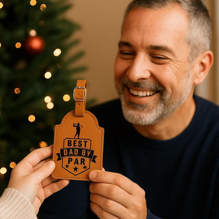 "Smiling man receives a tan leather golf bag tag engraved with 'Best Dad By Par' in black text and golf-themed design. The scene is warmly lit with a decorated Christmas tree in the background, creating a festive and heartfelt gift-giving moment."