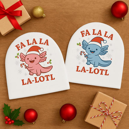 "Two white baby hats featuring festive axolotl designs are displayed on a wooden surface surrounded by Christmas decorations. One hat shows a pink axolotl wearing a Santa hat and holding a candy cane, and the other shows a blue axolotl with the same holiday design. Both hats include the phrase 'Fa La La La-Lotl' in red text. Around the hats are wrapped gifts, red ornaments, holly leaves, and pine cones."