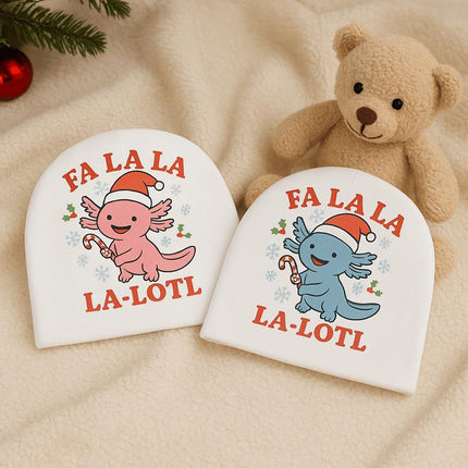 "Two white baby beanies featuring festive axolotl designs — one pink and one blue — each wearing a Santa hat and holding a candy cane, with the text 'Fa La La La-Lotl' printed in red. The hats are displayed on a soft cream blanket beside a tan teddy bear, evergreen branch, and red ornament, creating a cozy holiday nursery scene."