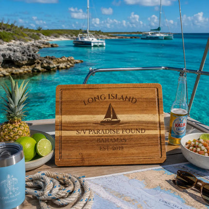 Long Island Bahamas sailboat cutting board displayed near a remote turquoise anchorage with rugged tropical coastline and distant sailboats in the southern Bahamas