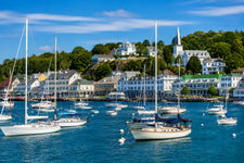 Sailboats anchored near Mackinac Island Harbor with historic waterfront buildings, lighthouse shoreline, and calm Great Lakes water under a bright summer sky.