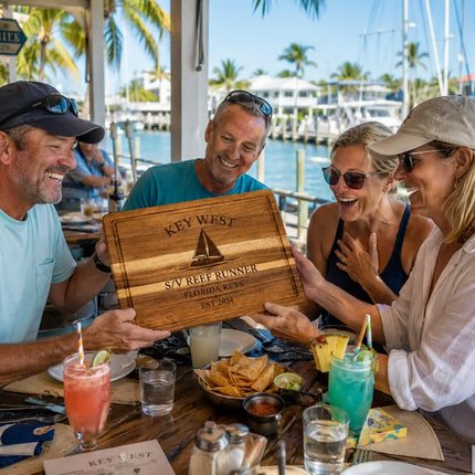 Friends celebrating at a Key West waterfront restaurant while admiring a personalized Harbor Collection cutting board commemorating a favorite harbor and sailing destination.